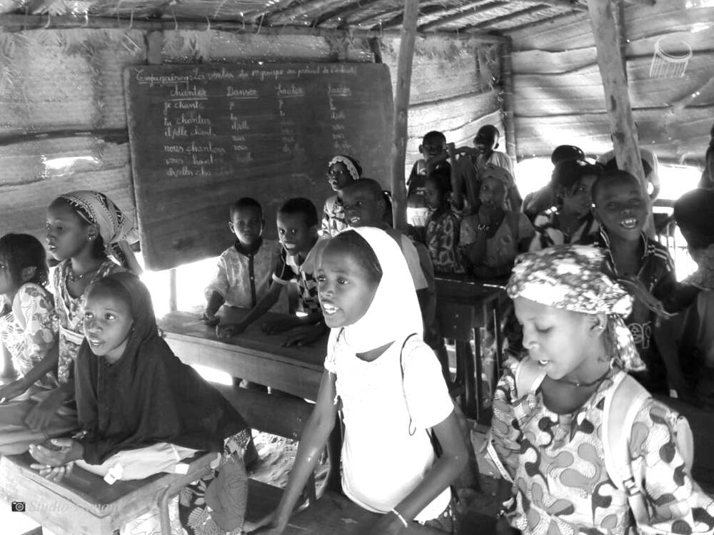 Des élèves assis sur des tables-bancs dans une classe abritée sous un hangar de paille et de bois, devant un tableau de conjugaison, au moment où la rentrée s’annonce difficile pour les enfants déplacés.