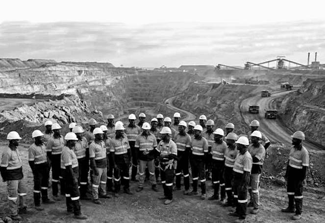 Une trentaine d'ouvriers en casque blanc et tenue à bandes réfléchissantes sont rassemblés en demi-cercle au bord d'une mine à ciel ouvert.
