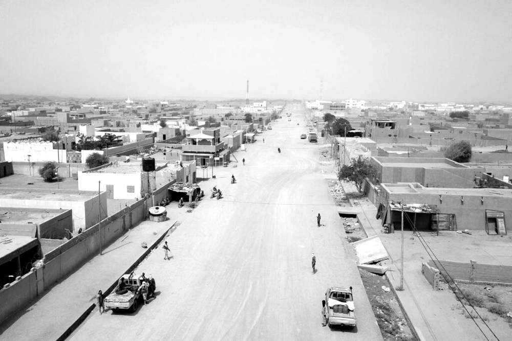 Une large avenue de Kidal vue en hauteur, bordée de maisons basses, où circulent quelques véhicules pick-up, des motos et des hommes armés à pied.