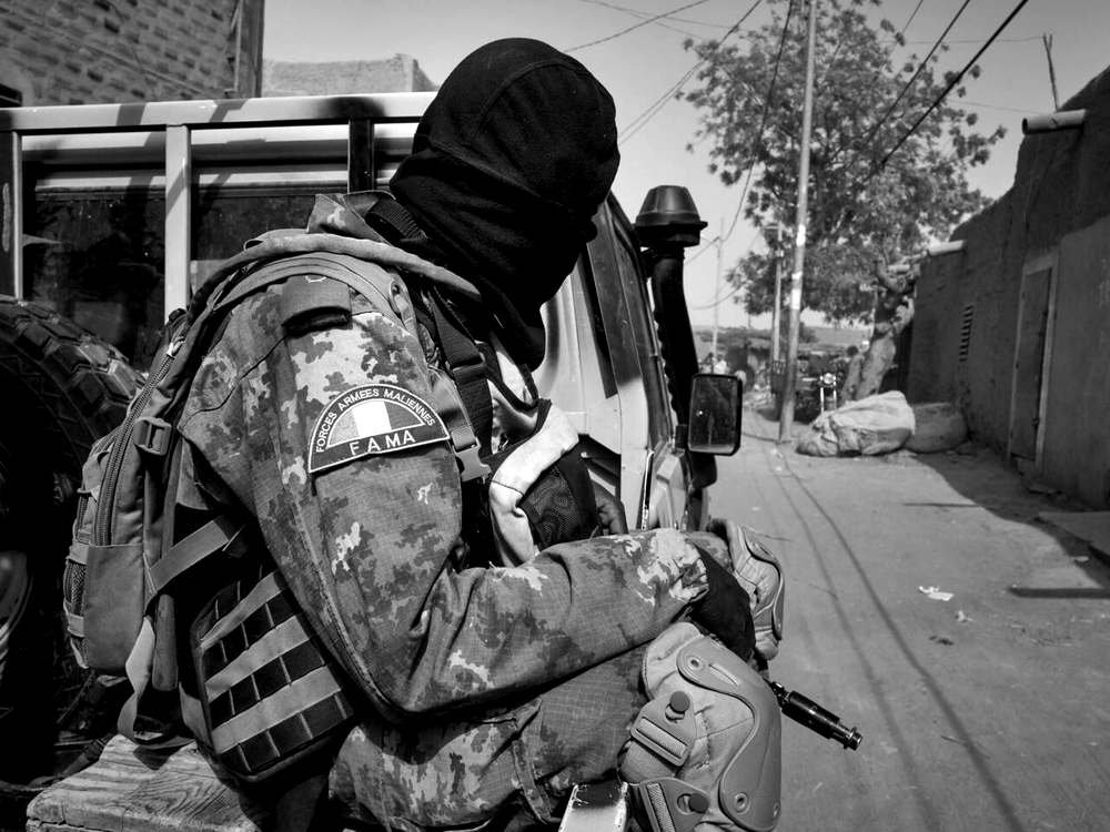 Un soldat des Forces armées maliennes, cagoulé et en tenue camouflée, assis à l’arrière d’un pick-up dans une ruelle bordée de murs en banco.