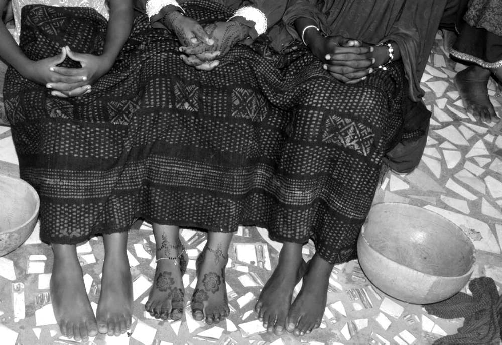 Trois jeunes femmes assises côte à côte, pagnes en bogolan, mains jointes et pieds ornés de henné, visages hors champ, sur un sol en carreaux cassés près de calebasses.