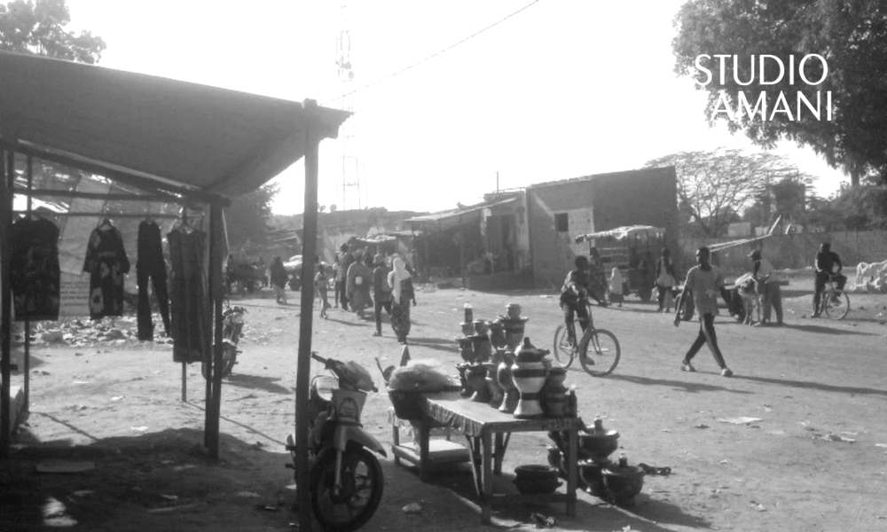 Une rue de marché animée, avec un étal de théières et d'ustensiles, des vêtements suspendus sous un hangar, des passants et des cyclistes, photographiée dans la localité de Tongolo coura, dans le cercle de Macina.