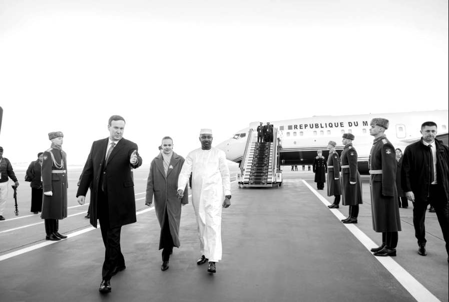 Un homme en boubou blanc et calotte, présenté par l'article comme le président de la Transition Assimi Goïta, marche sur le tarmac aux côtés d'officiels, devant une garde d'honneur et un avion marqué « République du Mali », à son arrivée à Moscou pour le sommet Russie–Afrique.